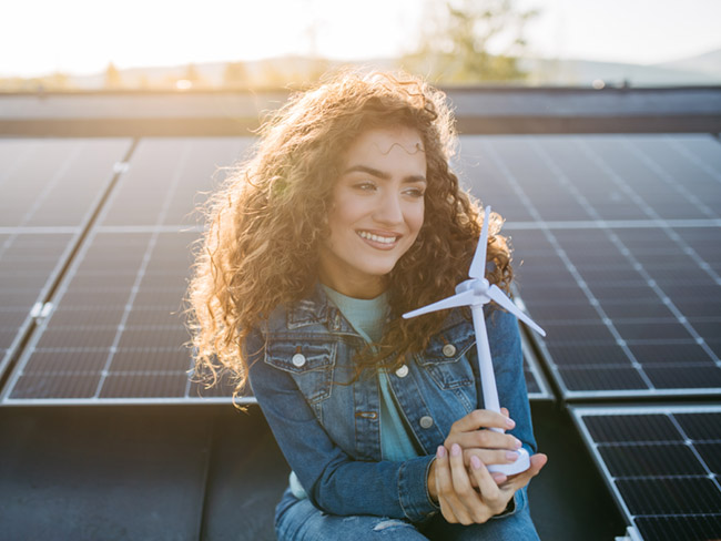 Portrait of young woman holding model of wind turbine on roof with solar panels.
